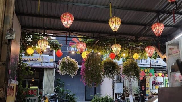 Colorful lanterns hang from a roof above red chairs and flowers at a street-side eatery.
