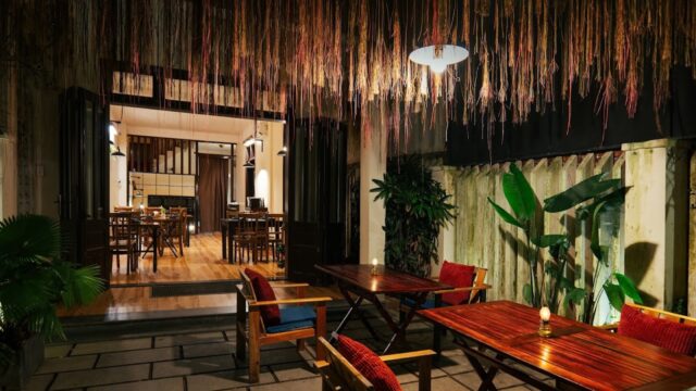 Cozy outdoor dining area with wooden tables, chairs, hanging decor, plants, and warm lighting at night.