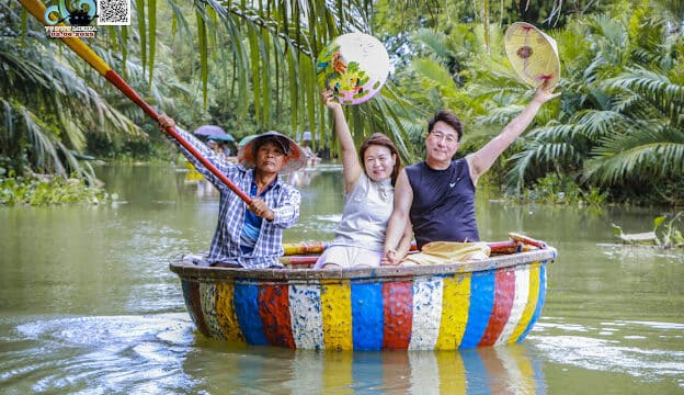 Three people smile and wave fans while riding a colorful basket boat on a lush Coconut Forest river.