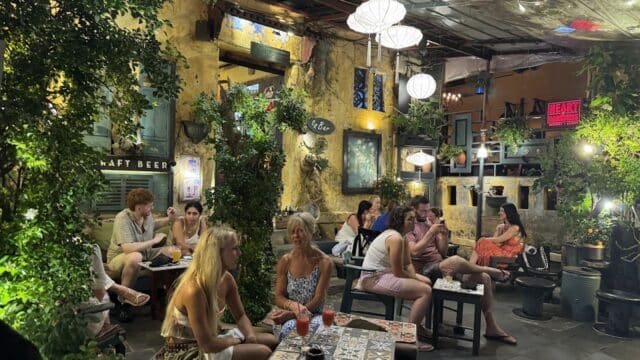 People sitting and chatting at tables in a cozy, greenery-filled Hoi An cafe with warm lighting and eclectic decor.