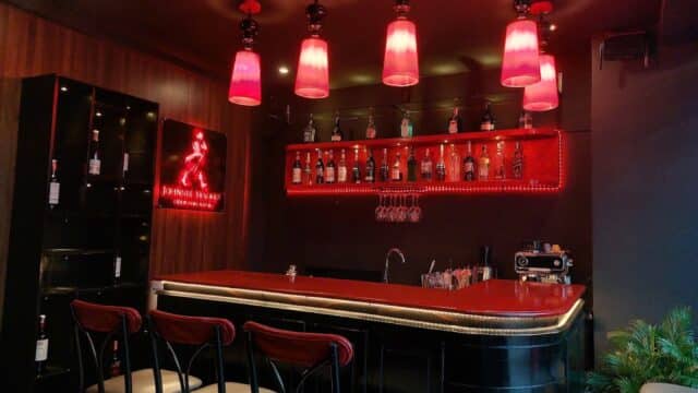 A modern bar in Hoi An with red lighting, bottles on display, bar stools, and a neon Johnnie Walker sign.
