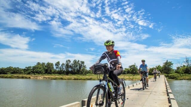 Three cyclists ride on a bridge over water under a bright blue sky with scattered clouds.