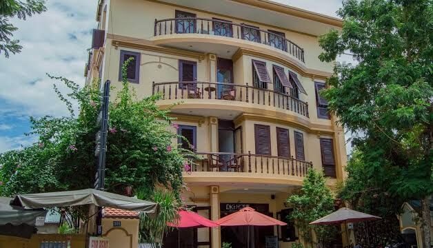 Four-story yellow DK's Backpacker Hotel with balconies, outdoor seating, red umbrellas, and lush greenery around.