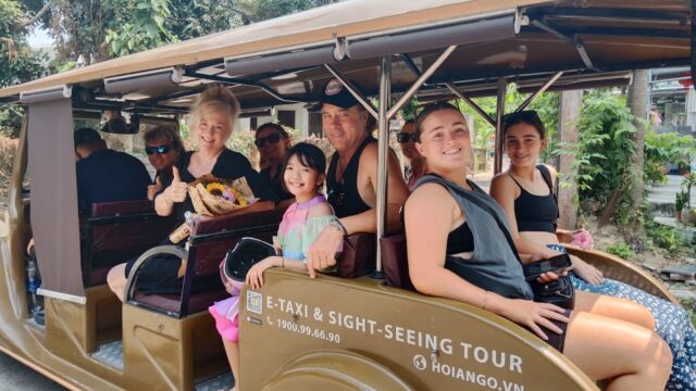 A group of smiling people sit in an open tour vehicle, ready for a sightseeing adventure in tropical Hoi An.