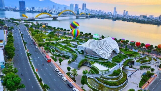 Aerial view of a riverside park, city skyline, and hot air balloons by a curved bridge at sunset with Dragon Travel Viet experiences.