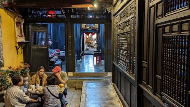Four people dine at a small table in Old House, savoring dumplings amid traditional wooden decor and intricate carvings.