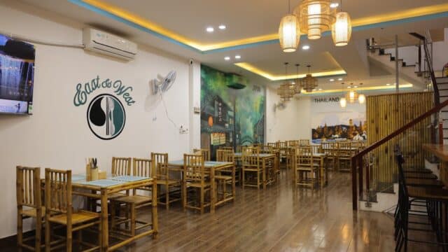 A modern Hoi An-inspired restaurant with bamboo furniture, wall art, and warm lighting, featuring an “East & West” sign.