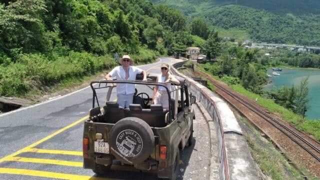 Two people stand in an open jeep on a mountain road, lush greenery and water behind them—an adventure with EasyRider’s Mr Thong.