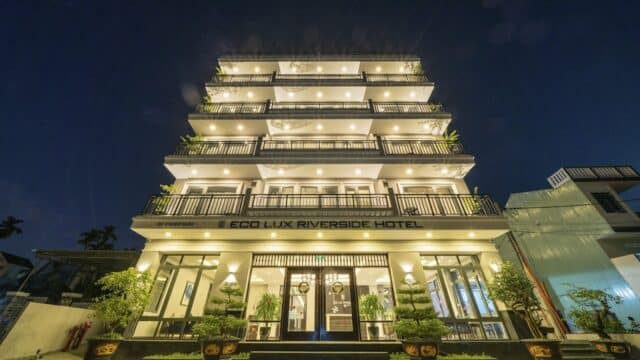 A brightly lit multi-story Eco Lux Riverside Hotel at night, with lush plants and trees welcoming guests at the entrance.