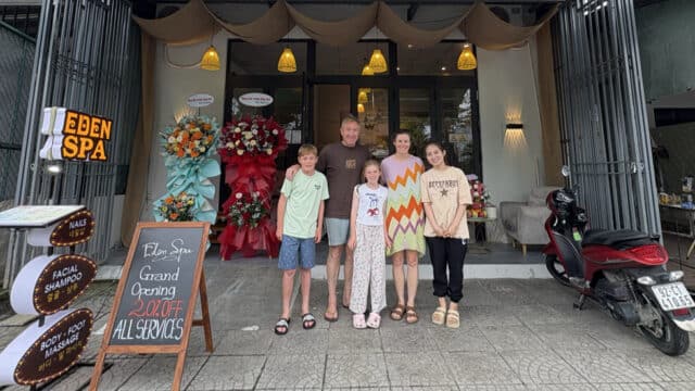 Five people stand smiling in front of a spa entrance with flower stands and a grand opening sign outside.