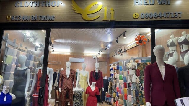 Clothing shop in Hoi An with mannequins in formal outfits, fabric rolls on shelves, and a sign reading eli above the entrance.