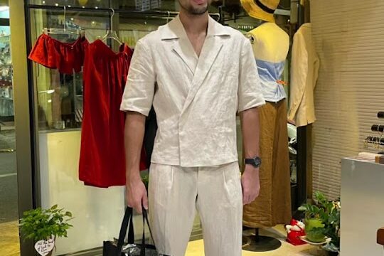 Man in a beige outfit smiling and holding a black shopping bag inside a Hoi An boutique with dresses on display.
