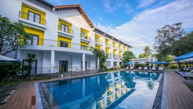 A modern Hoi An hotel with yellow accents overlooks a clear swimming pool surrounded by lounge chairs and trees.