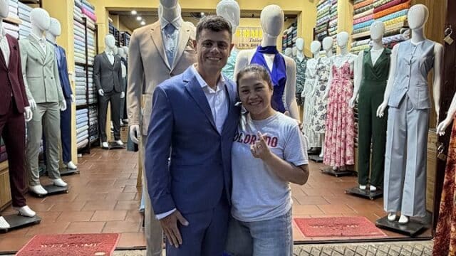 Two people smile and pose in front of a tailor shop, surrounded by mannequins dressed in suits and dresses.