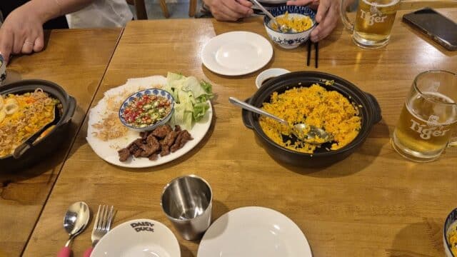 A wooden table set with plates of rice, grilled meat, vegetables, and mugs of beer, with people dining.