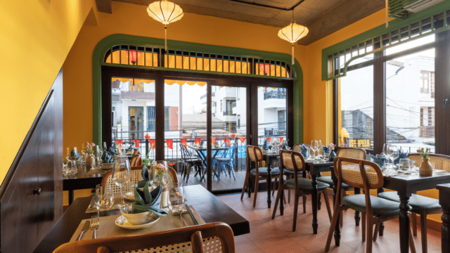 Faifoo Central Restaurant is a bright, cozy spot with set tables, cane chairs, and large windows opening to a balcony with city views.
