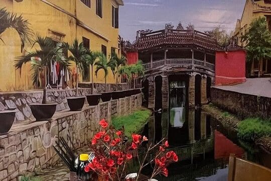A marble table with a vase of red flowers sits under a lantern, in front of a Hoi An mural of buildings and a bridge.
