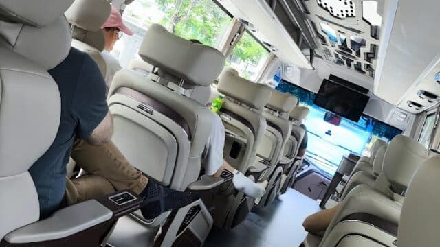 People are seated inside a modern Hoi An bus with large windows, beige seats, and blue lighting near the driver.