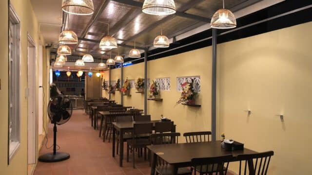 A cozy indoor dining area with wooden tables, chairs, hanging lights, and flower decorations on yellow walls.