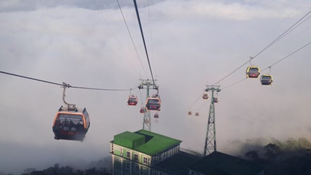 Cable cars glide above clouds and a green-roofed building, offering stunning views near the iconic Golden Bridge on the mountain.