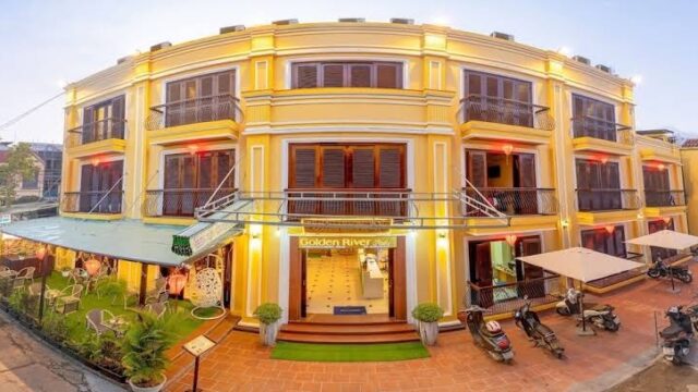 Golden River Hotel: Yellow three-story hotel with balconies and outdoor seating, motorbikes parked in front under a clear sky.