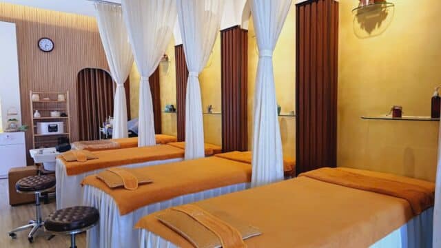 Three massage beds with orange covers and white curtains await in a serene, well-lit room at Goodnesss Spa, your haven for wellness.