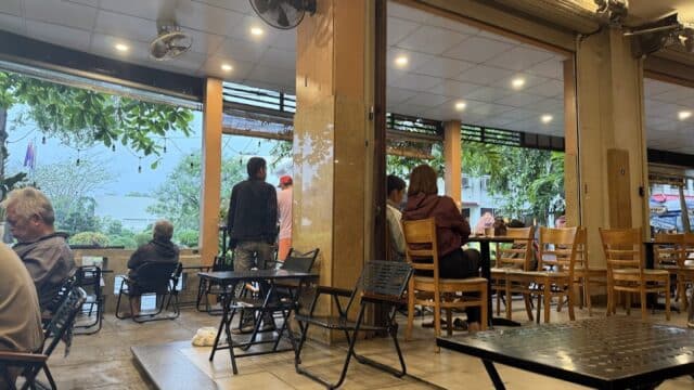 People sitting and standing in an open-air café with wooden chairs and tables in Hoi An, looking out toward lush greenery.