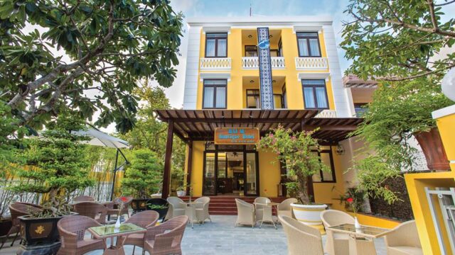 Hai Au Hotel is a yellow three-story hotel with outdoor seating, surrounded by trees and greenery under a blue sky.