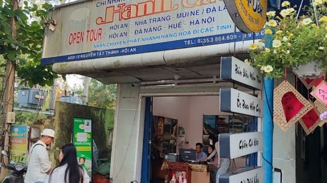 People stand outside a travel agency in Hoi An with a tour sign and a yellow bus stop sign on a city sidewalk.