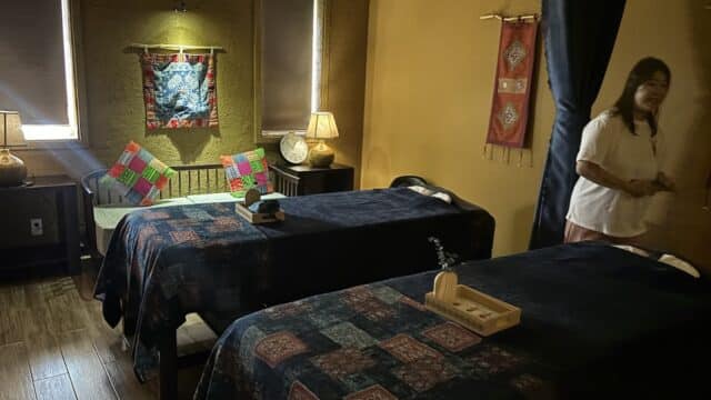 Dimly lit massage room with two beds, colorful decor, and a woman standing on the right side.