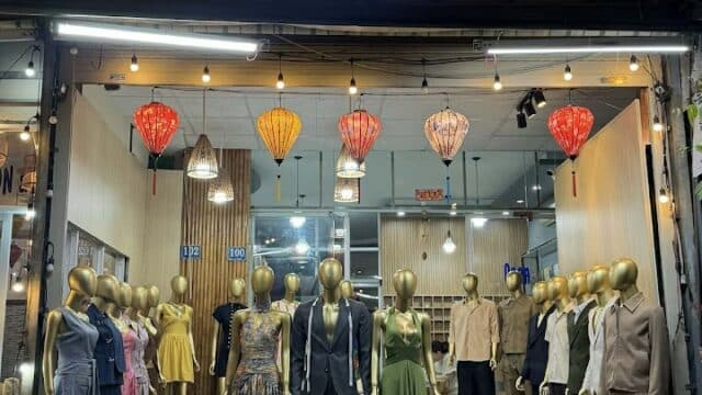 Clothing shop at night in Hoi An with mannequins in suits and dresses, colorful lanterns, and a sign reading Hey! Tailor.