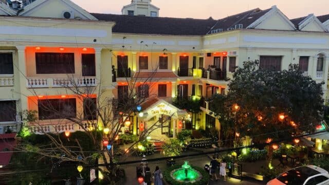 Hoa Binh Hostel features glowing lights, a green fountain, trees, parked cars, and people standing outside at dusk.