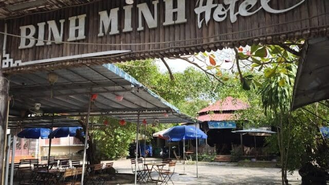 Entrance of Binh Minh Hotel in Hoi An with outdoor seating, trees, and blue umbrellas under a sunny sky.