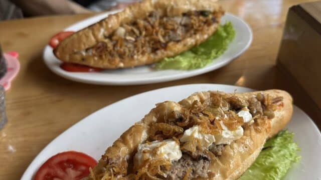 Two oval sandwiches with meat and toppings on white plates at Art Cafe, a cozy Hoi An Cafe, served with lettuce and tomato slices.