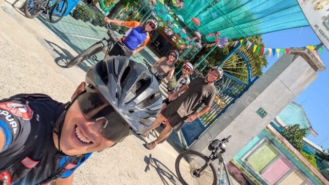 Five cyclists smile for a group selfie outside a colorful, decorated building in Hoi An on a sunny day.