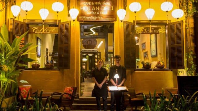 Two staff stand outside a warmly lit Hoi An restaurant entrance at night, surrounded by lanterns and lush plants.