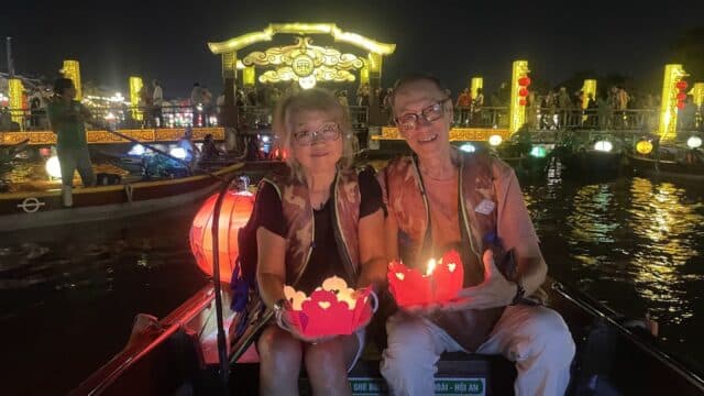 Two people sit in a boat at night on a Hoi An travel adventure, holding glowing lanterns with a brightly lit bridge behind them.