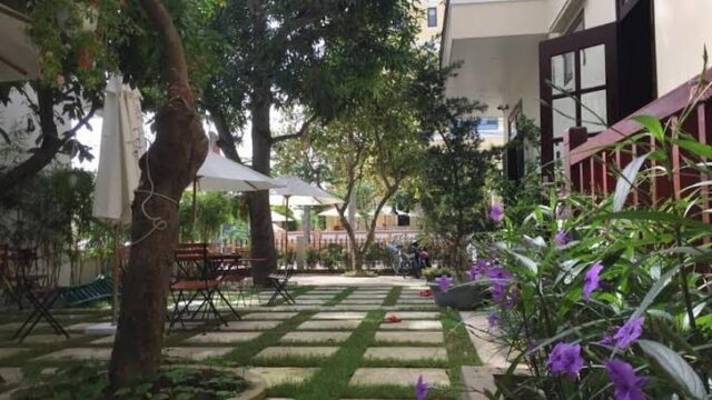 Garden with stone pathways, outdoor tables, chairs, umbrellas, and purple flowers beside a Hoi An heritage homestay.