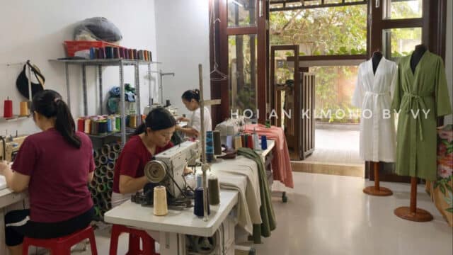 Three women sew at tables in a Hoi An tailor shop with spools of thread and kimonos displayed on mannequins.