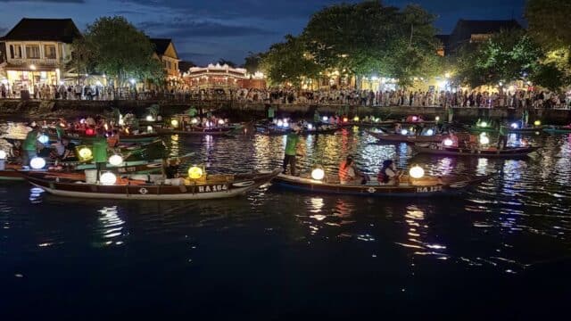 Boats decorated with colorful lanterns float on the river at night in Hoi An, as crowds enjoy the Lantern Boat Tour from the riverside.