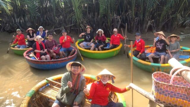 A group enjoys a local eco tour in Hoi An, smiling in round basket boats on a brown river amidst lush green palm trees.