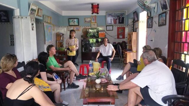 A group of people sit and talk in a brightly lit living room, sharing stories about their recent Hoi An Tours amidst photos and certificates.