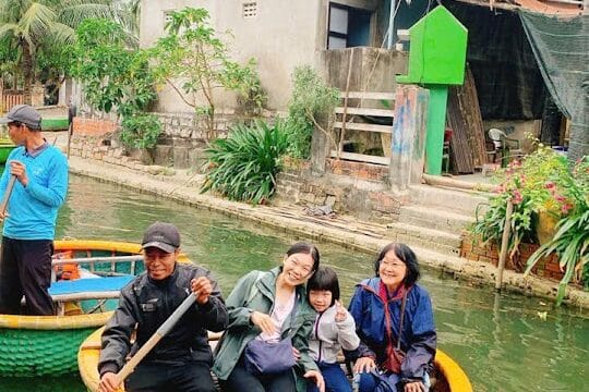 Four people sit in a round basket boat on a canal in Hoi An, with houses and greenery visible in the background.