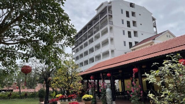 A garden with potted plants and flowers beside a long River Town building with red roof and a tall white structure behind.