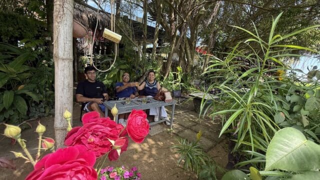 Three people sit on a bench in a lush Hoi An garden with red roses, perhaps resting after a vibrant shopping tour.