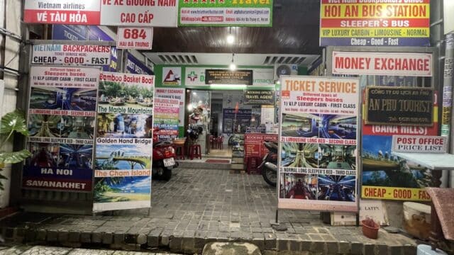 Travel agency storefront in Hoi An, Vietnam, covered with colorful signs for tours, buses, money exchange, and travel services.