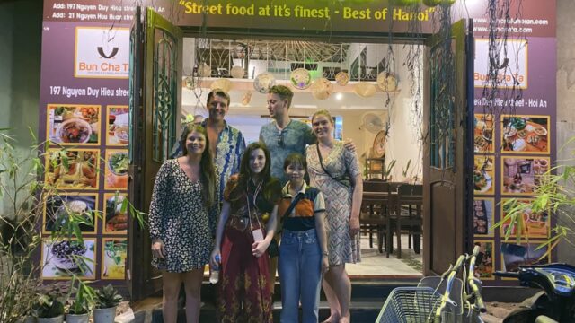 Six people pose and smile in front of a yellow-lit Vietnamese restaurant named Bun Cha Ta at night, ready for Street Food Tours.