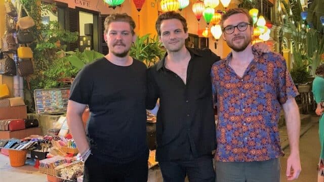 Three men pose together on a colorful Hoi An street with hanging lanterns, a vibrant market, and tempting street food in the background at night.