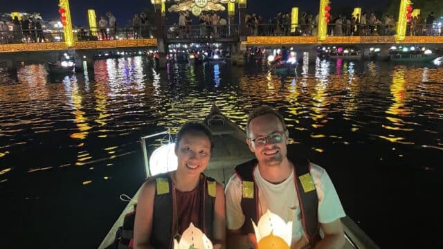 A smiling couple in a boat at night on a Hội An Tour, holding lanterns with a brightly lit bridge behind them.