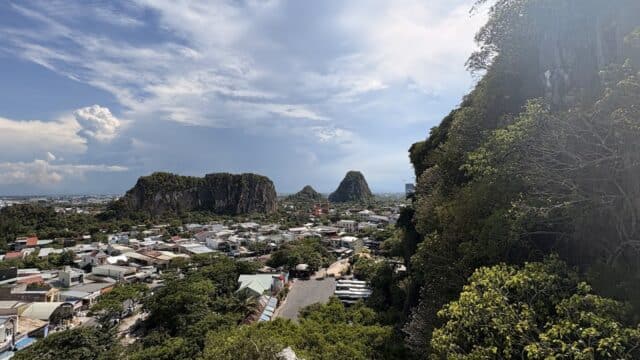 A panoramic view of a town along the Hoi An Trail, with green hills, rocky mountains, and a partly cloudy sky in the background.